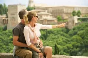 Middle aged couple sitting on a stone wall pointing towards the Alhambra in Granada Spain during a sightseeing tour