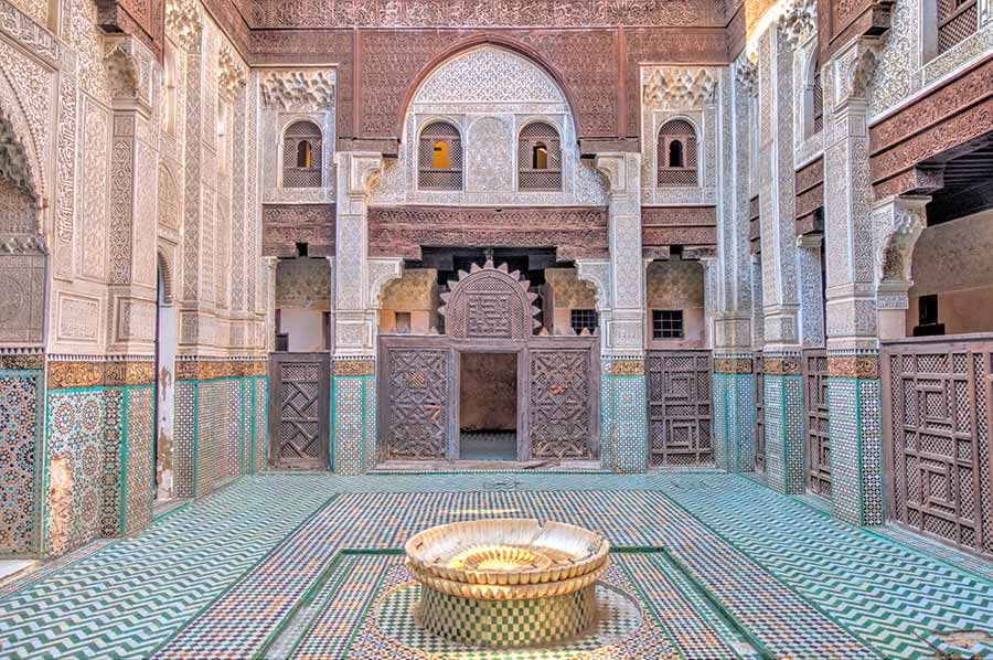 Intricate interior courtyard of Bou Inania Madrasa in Fes, Morocco showcasing Islamic architecture, ornate tilework, carved wood, and a central fountain