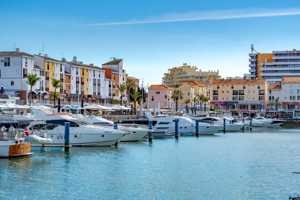 Luxury yachts docked at Vilamoura Marina in the Algarve, Portugal, with colorful waterfront buildings and palm trees under a clear blue sky