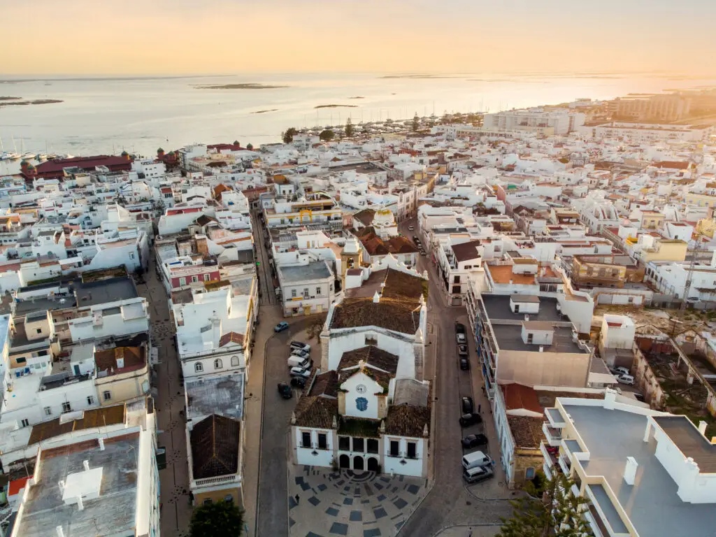 Aerial view of Olhão town with a historic church in the foreground, whitewashed buildings, marina, and Ria Formosa lagoon at sunset in Algarve, Portugal
