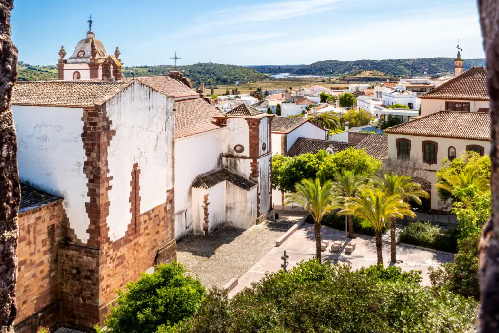 View from Silves Castle overlooking Silves Cathedral, the town square with palm trees, and the rooftops of Cidade de Silves leading to the Arade River under a sunny sky in Algarve, Portugal