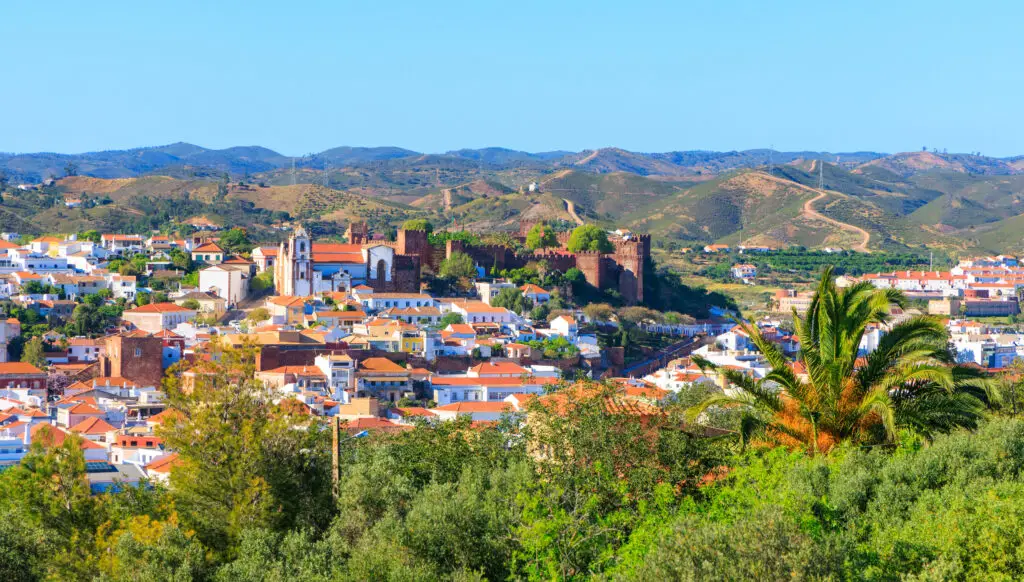 Panoramic view of Silves city in the Algarve region of Portugal featuring the historic Silves Castle, whitewashed houses with orange rooftops, and rolling green hills in the background.
