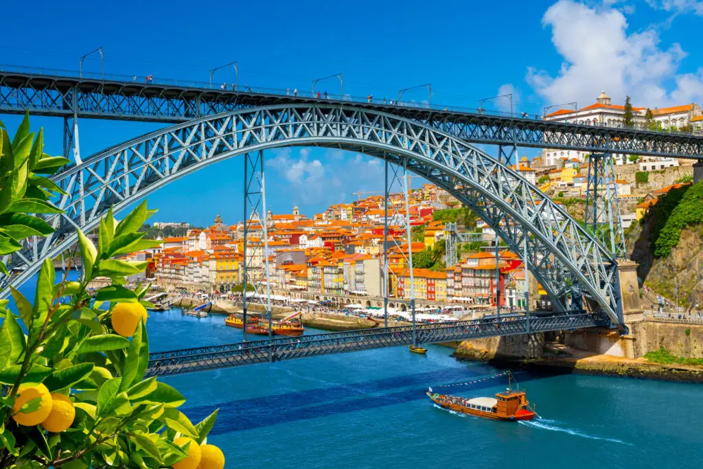 Colorful summer view of Porto, Portugal featuring the Dom Luís I Bridge over the Douro River, traditional boats, and the historic Ribeira district under a bright blue sky
