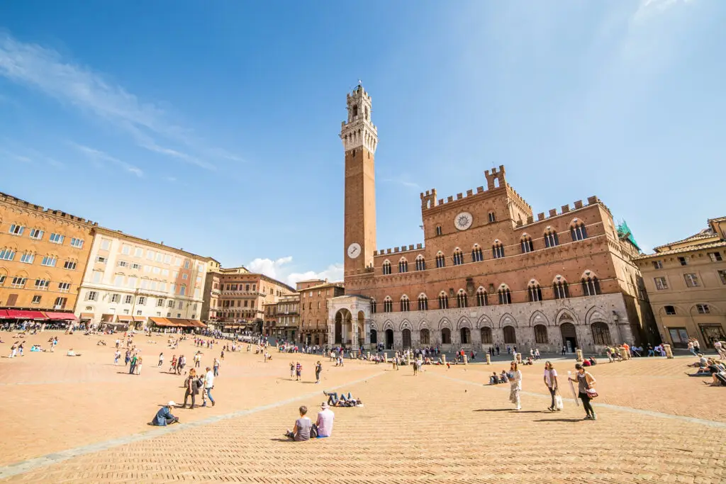 Tourists relaxing and exploring Piazza del Campo in Siena Italy with the historic Palazzo Pubblico and Torre del Mangia under a bright blue sky