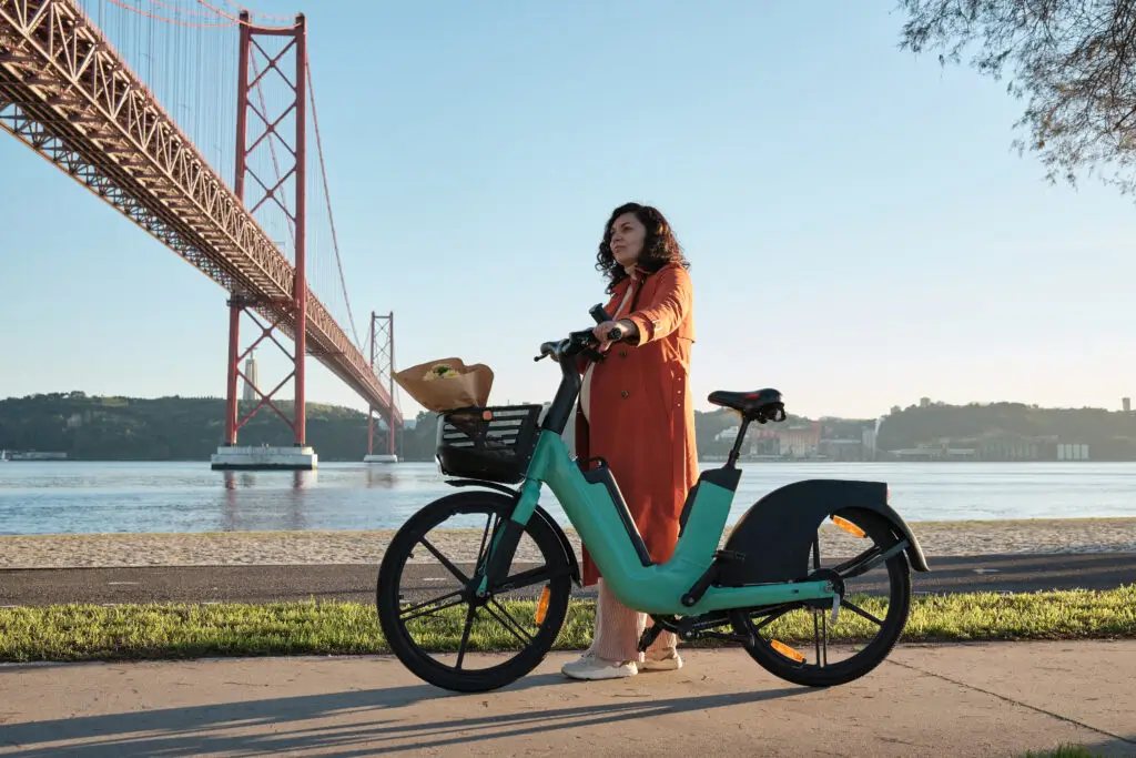Pregnant woman in orange coat standing with an electric bicycle on the Lisbon promenade near the 25 de Abril Bridge in Portugal