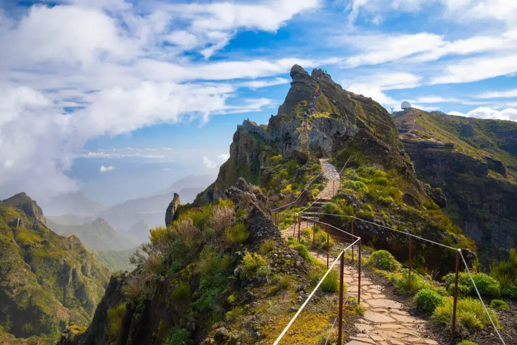 Scenic mountain trail leading to the summit of Pico do Arieiro on Madeira Island, Portugal, with stunning views of rugged cliffs, lush greenery, and dramatic clouds above the Atlantic Ocean